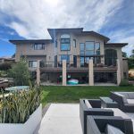 Backyard of a modern beige house with large glass windows and string lights on a sunny day.