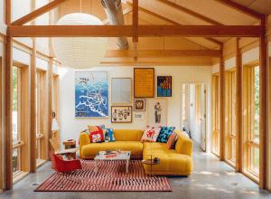 Sunlit living room with a bright yellow sectional sofa, colorful quilted cushions, and a wall gallery of framed art pieces under wooden beams.