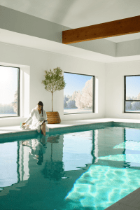 Woman in a white robe sits at the pool edge, reading a magazine beside a potted tree indoors. Snowy trees visible through large windows behind her.