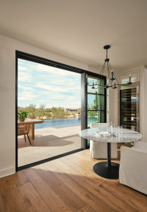 Interior dining area with a hanging black chandelier, round marble table, and white chairs; large sliding doors open to a pool patio outside.