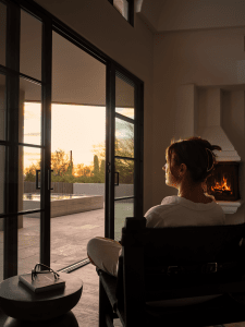 Person seated in a chair by large glass doors watching a sunset, with a lit fireplace on the right and a notebook with glasses on a side table nearby.