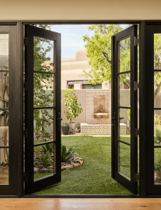 Open black-framed glass doors lead to a sunny courtyard with a tiered stone fountain, greenery, and beige stucco walls.