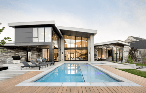 Modern house with large glass windows, stone facade, and an in-ground swimming pool surrounded by a wooden deck.