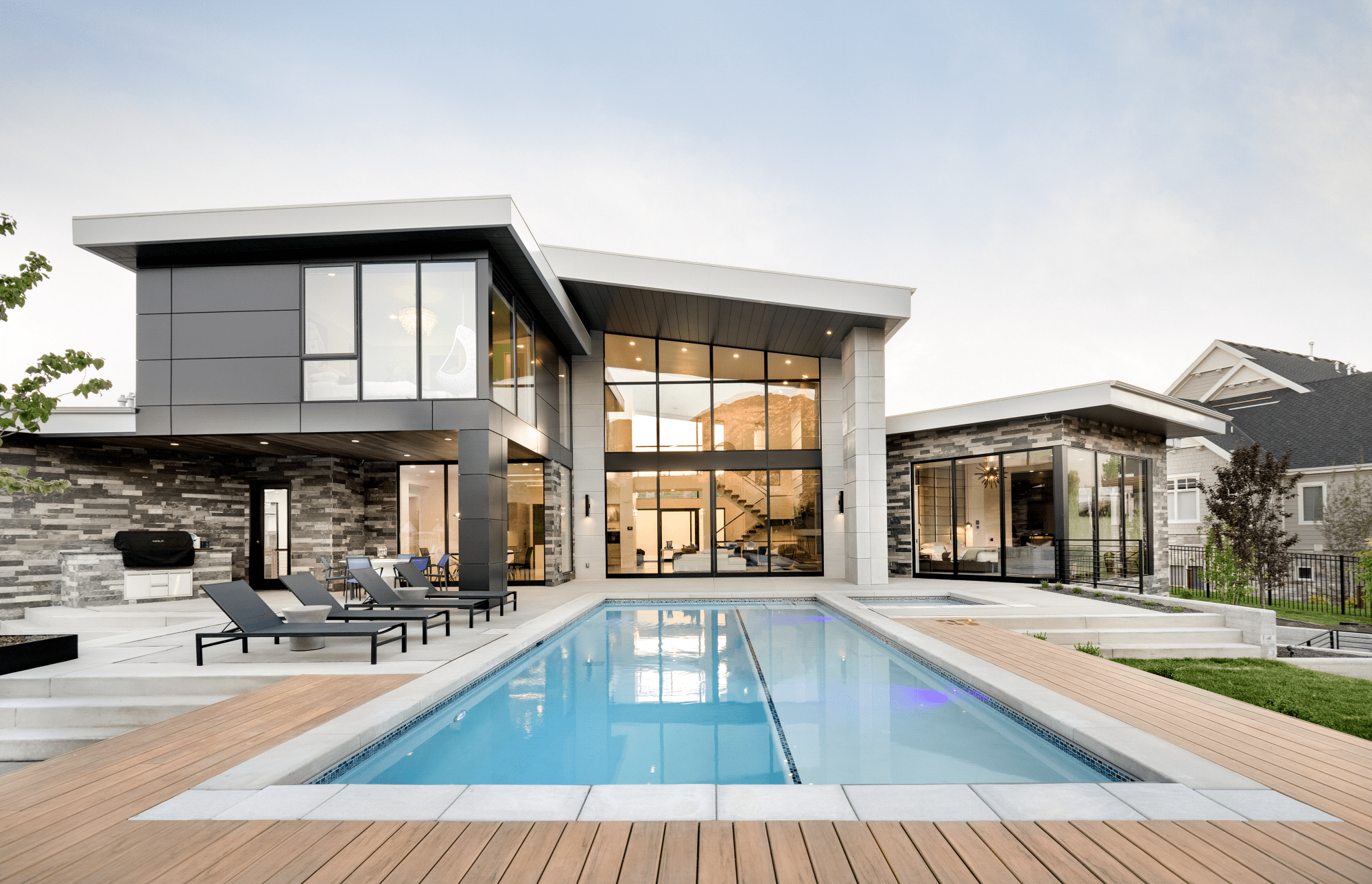 Modern house with large glass windows, stone facade, and an in-ground swimming pool surrounded by a wooden deck.
