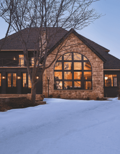 Stone house with a large arched window, warm interior lights, and snow on the front yard; bare tree branches in the foreground.