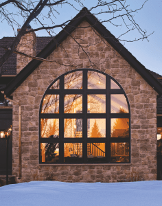 Stone house facade with a large arched window reflecting a sunset, framed by leafless tree branches and a snow-covered foreground