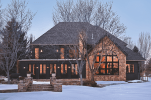 Winter scene of a dark-toned, stone-and-wood house with large windows and leafless trees in front; snow on the ground.
