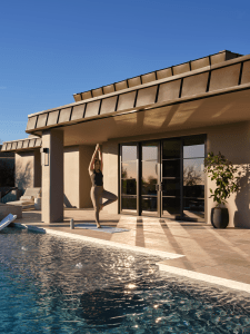Woman practicing yoga in tree pose on a mat beside a swimming pool in front of a modern beige house with large glass doors