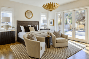 Bright, contemporary bedroom with a dark wood bed, neutral bedding, and two beige striped accent chairs by a coffee table near large French doors to a sunny backyard.