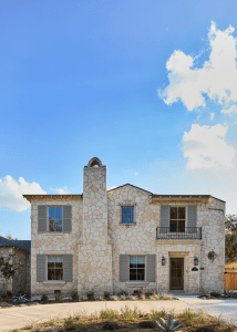 Stone house with a stone facade, gray shutters, and a wooden front door under a small balcony against a bright blue sky.