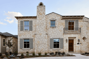 Stone townhouse facade with beige stonework, gray shutters, and multiple windows on two stories, including a small wrought-iron balcony near the center-right.