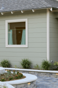 Exterior view of a pale green house with a white-framed window and turquoise curtains, with a curved concrete planter and green plants in front.