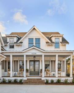 White two-story house with a covered front porch, gray shutters, decorative gables, and the number 4206; patio chairs and potted plants on the porch.