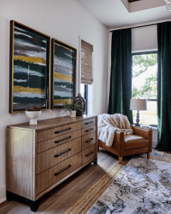 Wooden dresser against a wall with two gold-framed abstract paintings, small plants, and a fur throw by a leather chair near a window with green curtains.