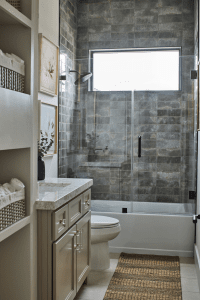 Modern bathroom with a glass-enclosed shower, gray tile walls, and a beige vanity with marble countertop and framed botanical art on the wall.