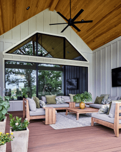 Covered outdoor living area with sectional sofa, wooden coffee tables, and green throw pillows under a wooden vaulted ceiling and black ceiling fan.
