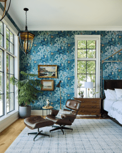 Corner of a bright living room with blue floral wallpaper, leather lounge chair and matching ottoman, and a windowed wall.