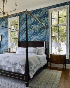 Elegant bedroom with a dark four-poster bed, blue floral wallpaper, and large windows overlooking trees and water outside.