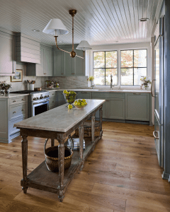 Bright kitchen with pale blue-green cabinets, marble countertops, and a rustic wooden island under a copper chandelier; large window fills the space with light.