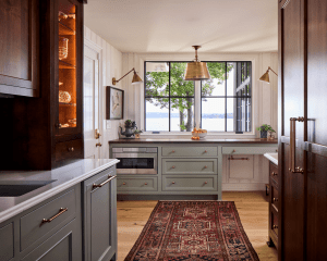 Kitchen with blue-gray cabinets, brass hardware, and a window view of trees and a lake; warm pendant lighting and a red patterned rug.