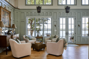Sunlit living room with cream upholstered armchairs arranged around a round wicker-topped coffee table, sage paneled walls, and large windows showing a lake view.