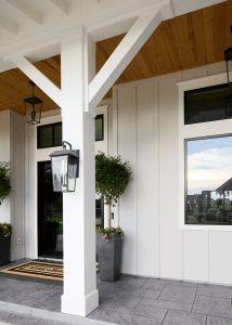 White front porch with a tall white column, black lanterns, a glass entry door, large window, and tall planters with greenery.