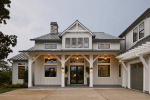Front view of a modern farmhouse with gray vertical siding, a metal roof, central dormer window, and lit lanterns on the covered porch.