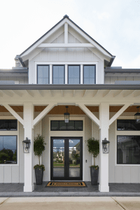 White modern farmhouse-style entrance with black-framed glass doors, tall white columns, and potted trees on either side of the doorway