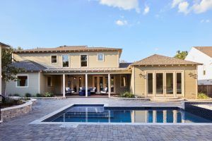 Backyard of a beige two-story house with a covered patio and rectangular pool, under a clear blue sky.