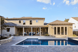 Backyard with a rectangular in-ground pool, stone patio, and a beige two-story house with a covered seating area.