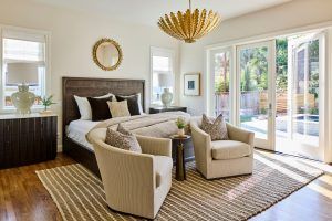 Bright master bedroom with a dark wood headboard, multiple decorative pillows, and beige upholstered chairs around a small table.