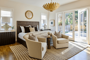 Bright bedroom with a dark wood bed, beige seating, and a woven rug; large glass doors open to a sunny backyard.