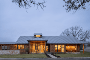 Modern brick house with large glass windows glowing from warm interior lights at dusk; metal roof and bare trees frame the scene.