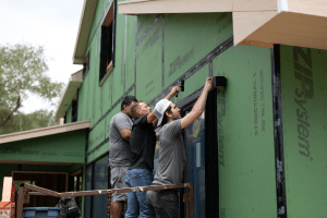 Group of construction workers installing a large glass storefront door on a green, under-construction exterior.