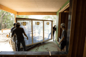 Construction workers install a large glass storefront window inside a wooden-framed building under construction.