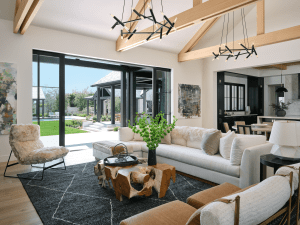 Open living area with floor-to-ceiling glass doors, exposed wooden beams, and a modern sculptural black chandelier above a natural wood coffee table and white sofa set.