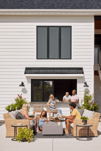People enjoying an outdoor gathering on a patio in front of a white house with horizontal siding and large central window, wicker seating, and string of lights (people chat and share drinks).
