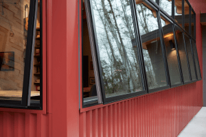 Exterior view of a red metal-clad building with tall angled glass windows reflecting bare trees outside