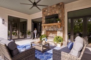 Outdoor covered patio with wicker seating, blue patterned rug, and stone fireplace; a TV mounted above the mantel and potted plants nearby.