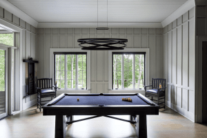 Pool table in a bright paneled room with a black circular chandelier and large windows showing greenery outside.