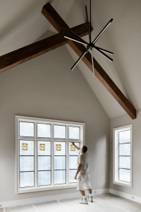 Worker in casual clothes stands by tall grid windows in a room with exposed wooden ceiling beams and a modern starburst chandelier overhead of the vaulted ceiling.