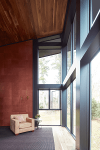 Cozy seating area by large floor-to-ceiling windows with a wooden ceiling and red-t tiled wall, outdoors visible through the glass.