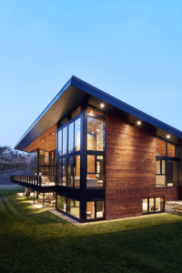 Modern, angular house with wood siding and expansive glass walls lit from within at dusk.