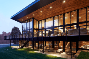Modern glass-walled building with warm wooden ceiling and outdoor seating on a lit balcony at dusk/sunset.