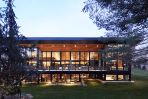 Modern glass-walled house with wood panels, warm interior lighting, and a front terrace surrounded by trees.