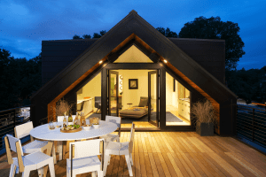 Outdoor wooden deck with a white dining table set for a meal; triangular glass entry and warm interior lights visible inside the house at dusk.