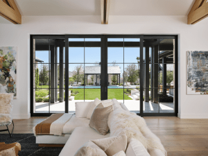 Living room with large black-framed glass doors opening to a sunny backyard and pool; beige sofa and fluffy throws in foreground.