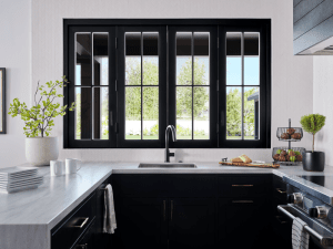 Modern kitchen with a large black-framed window over a white marble island and sink, plants on the left and a fruit basket on the right.