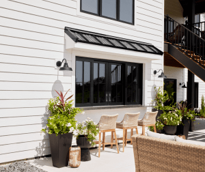 Outdoor patio area along a white house with black-framed doors, wicker chairs, and large planters with greenery