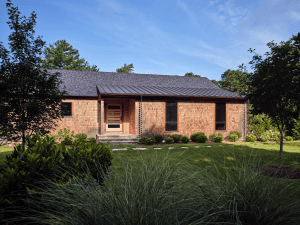 Brick single-story house with a front porch, surrounded by lush landscaping and trees under a clear blue sky.